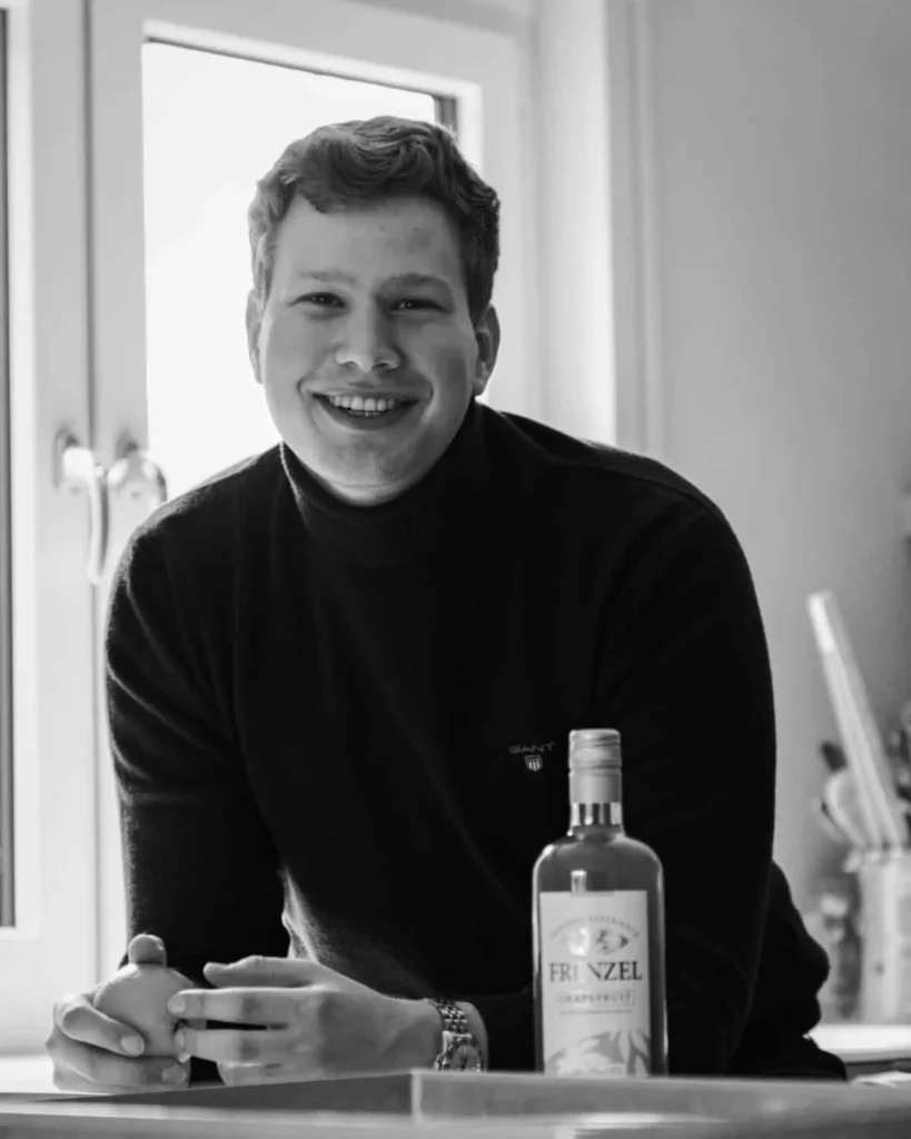 Johannes M. Frenzel, Gründer von FRENZEL Grapefruit, lächelt in die Kamera. Vor ihm auf dem Tisch steht eine Flasche FRENZEL Grapefruit Likör, in seinen Händen hält er eine frische Grapefruit.